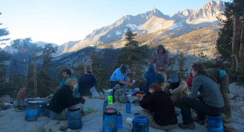 People sit amid their gear on rocky ground. There are rocky mountains in the background. 